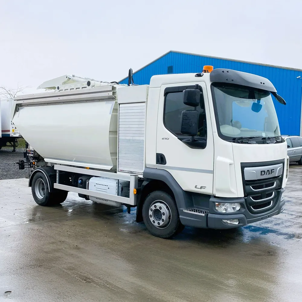 White DAF refuse truck parked on wet concrete in front of a blue industrial building.
