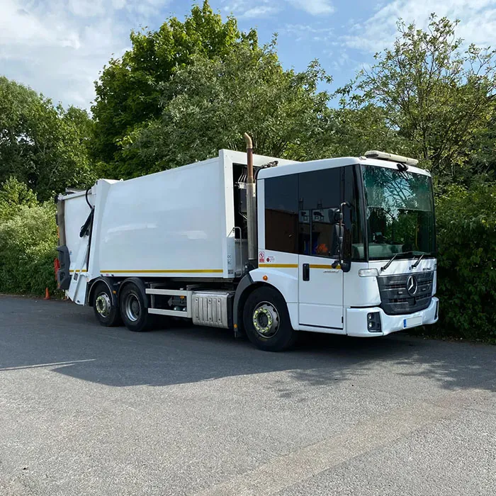 White Mercedes-Benz refuse collection truck parked on a road beside green trees under a partly cloudy sky.