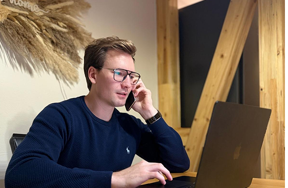Junger Mann mit Brille spricht am Telefon und arbeitet an einem Laptop an einem Holztisch.