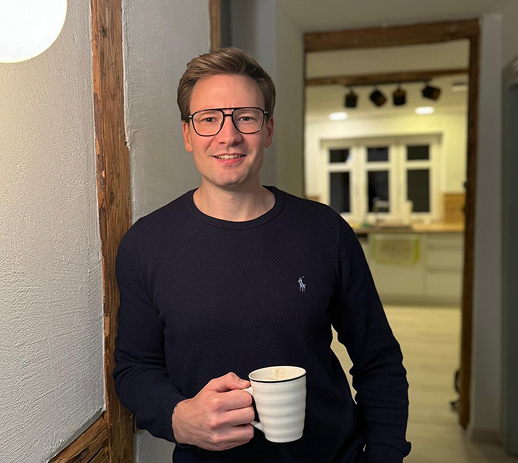 Ein lächelnder Mann mit Brille steht in einem Hausflur und hält eine weiße Kaffeetasse.