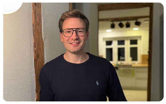 Mann mit Brille und dunkelblauem Pullover steht vor einer weißen Wand und lächelt, im Hintergrund eine helle Küche.