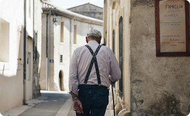 Ein älterer Mann mit grauem Haar und Hosenträgern geht eine enge Gasse zwischen historischen Gebäuden entlang.