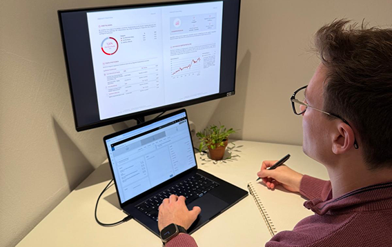 Person sitzt an einem Schreibtisch, arbeitet an Laptop und macht Notizen, während auf einem oberen Bildschirm Diagramme und Daten angezeigt werden.
