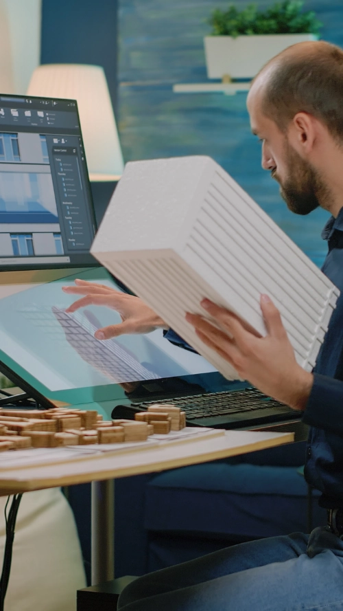Person working on a 3D interior design software showing a living room layout on a computer monitor in a bright office.