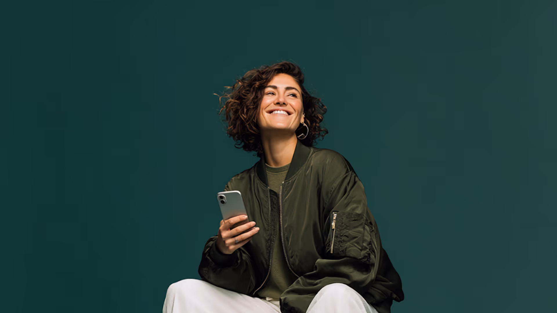 Smiling woman with curly hair wearing a green jacket and holding a smartphone against a dark green background.