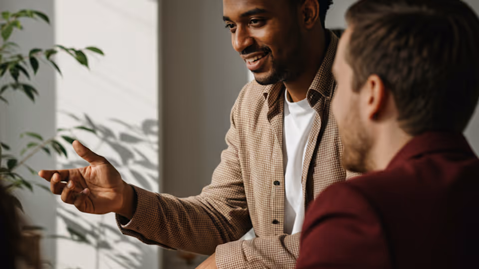 Smiling man in checkered shirt gesturing while talking to another man in a maroon shirt indoors.
