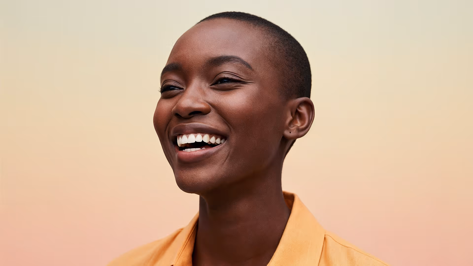Smiling person with a shaved head wearing a yellow shirt against a light gradient background.