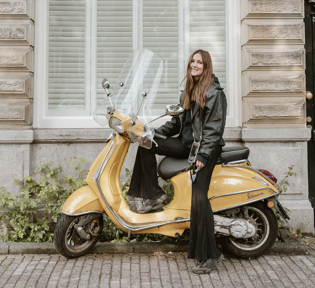 Woman in black leather jacket sitting on a yellow Vespa scooter parked by a building with white shutters.