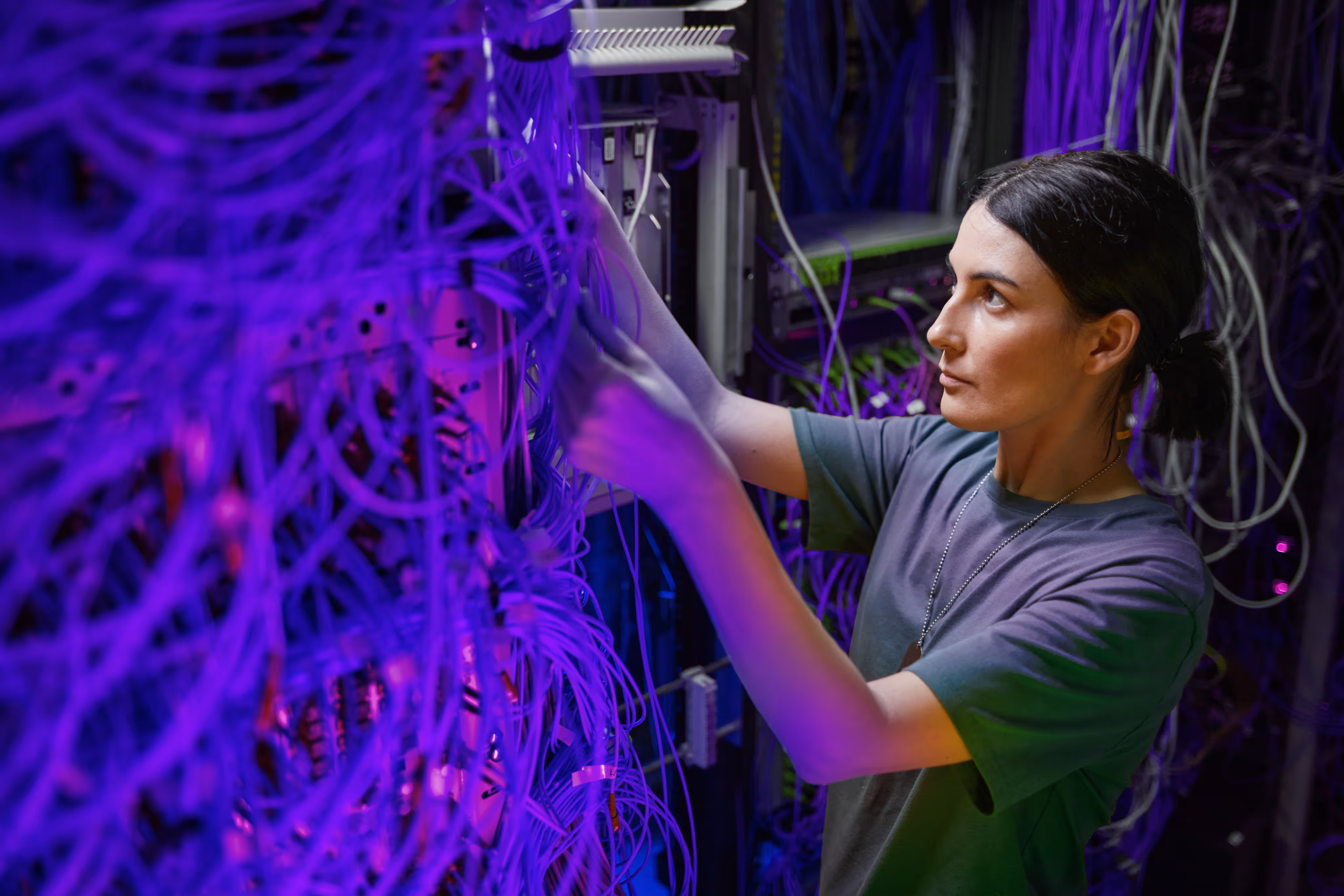 Woman working on network cables and servers in a dimly lit data center with purple lighting.