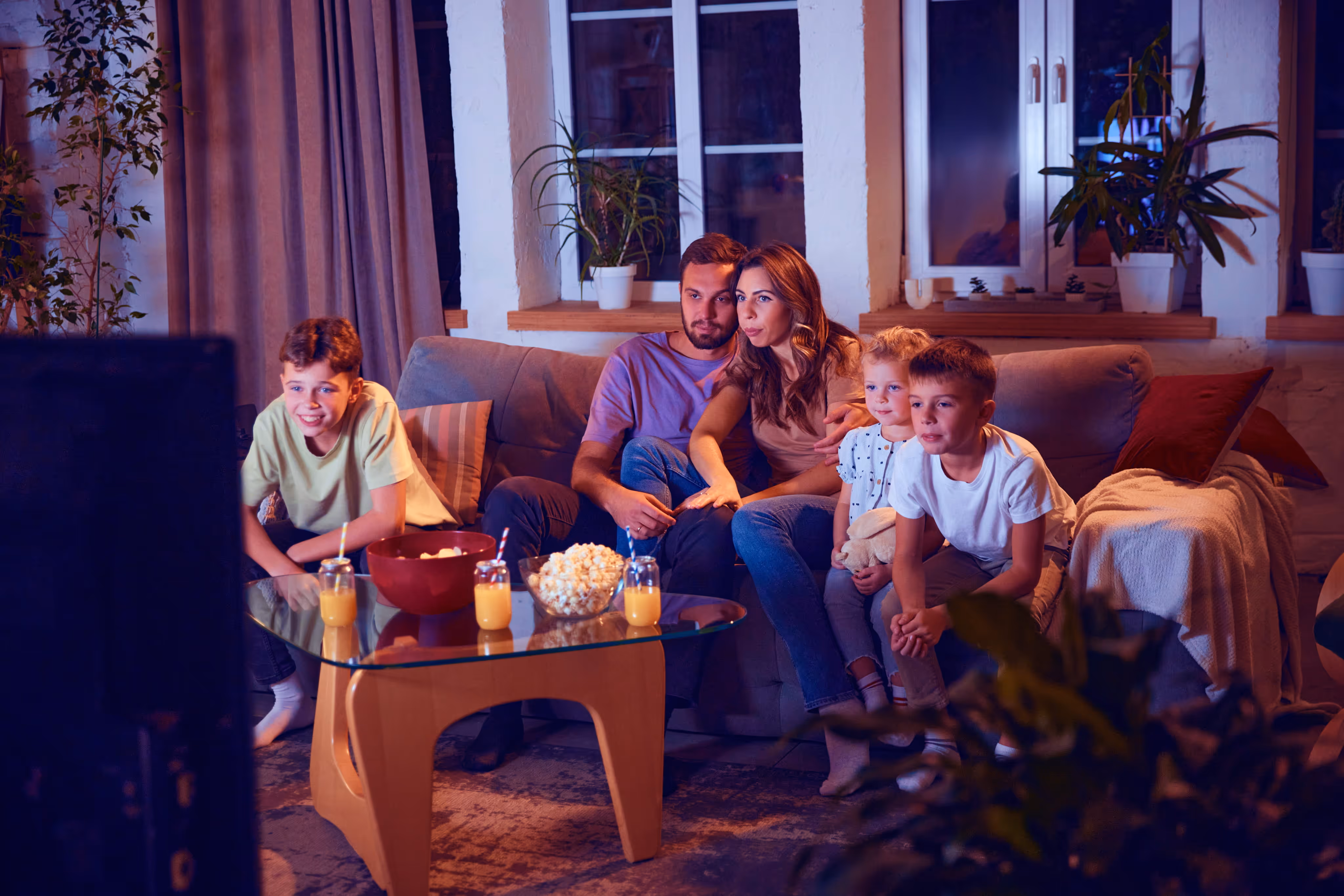 Family of five watching TV together in a cozy living room with snacks on the glass table.