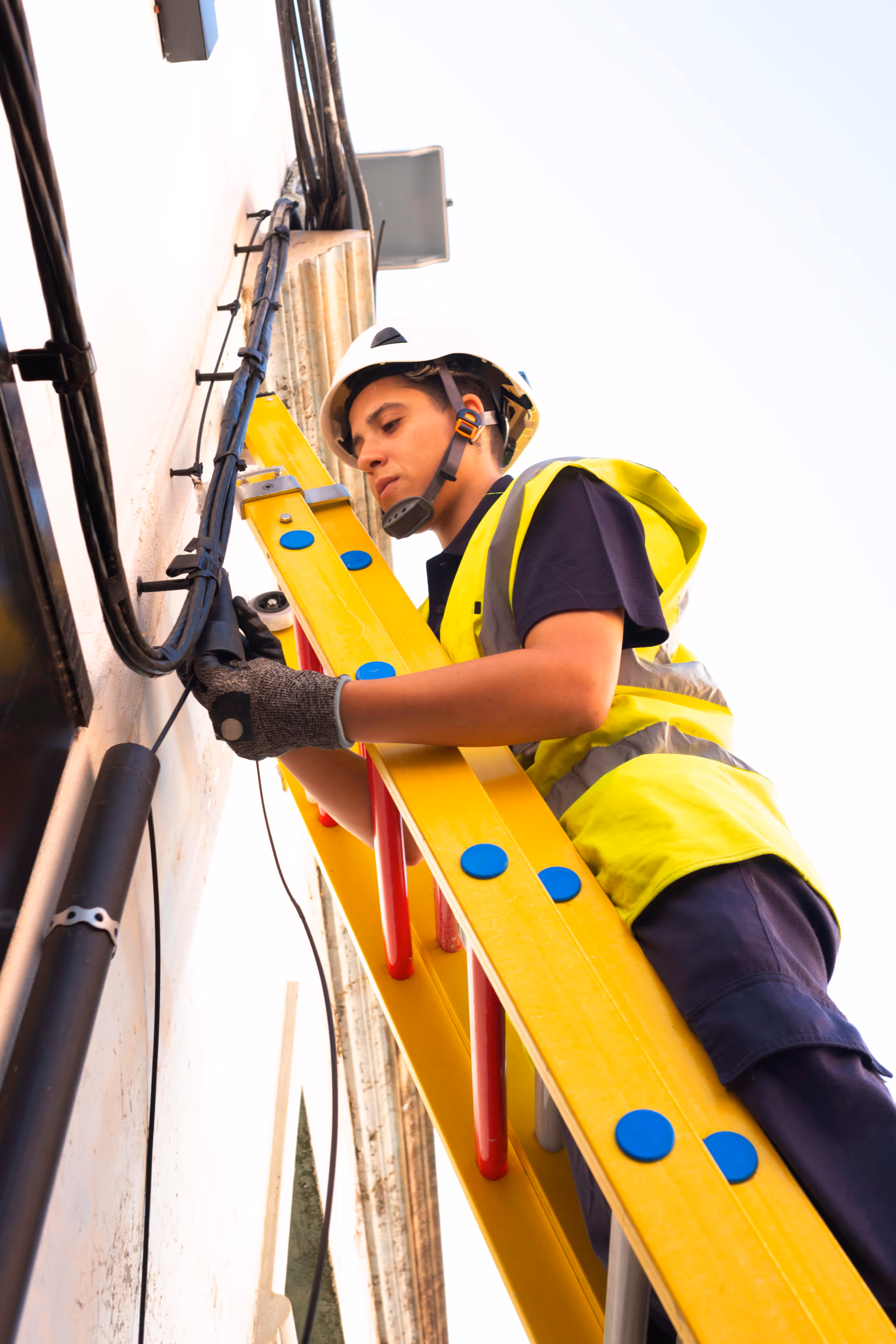 Electrician in a helmet and vest on a yellow ladder working on cables attached to a wall.