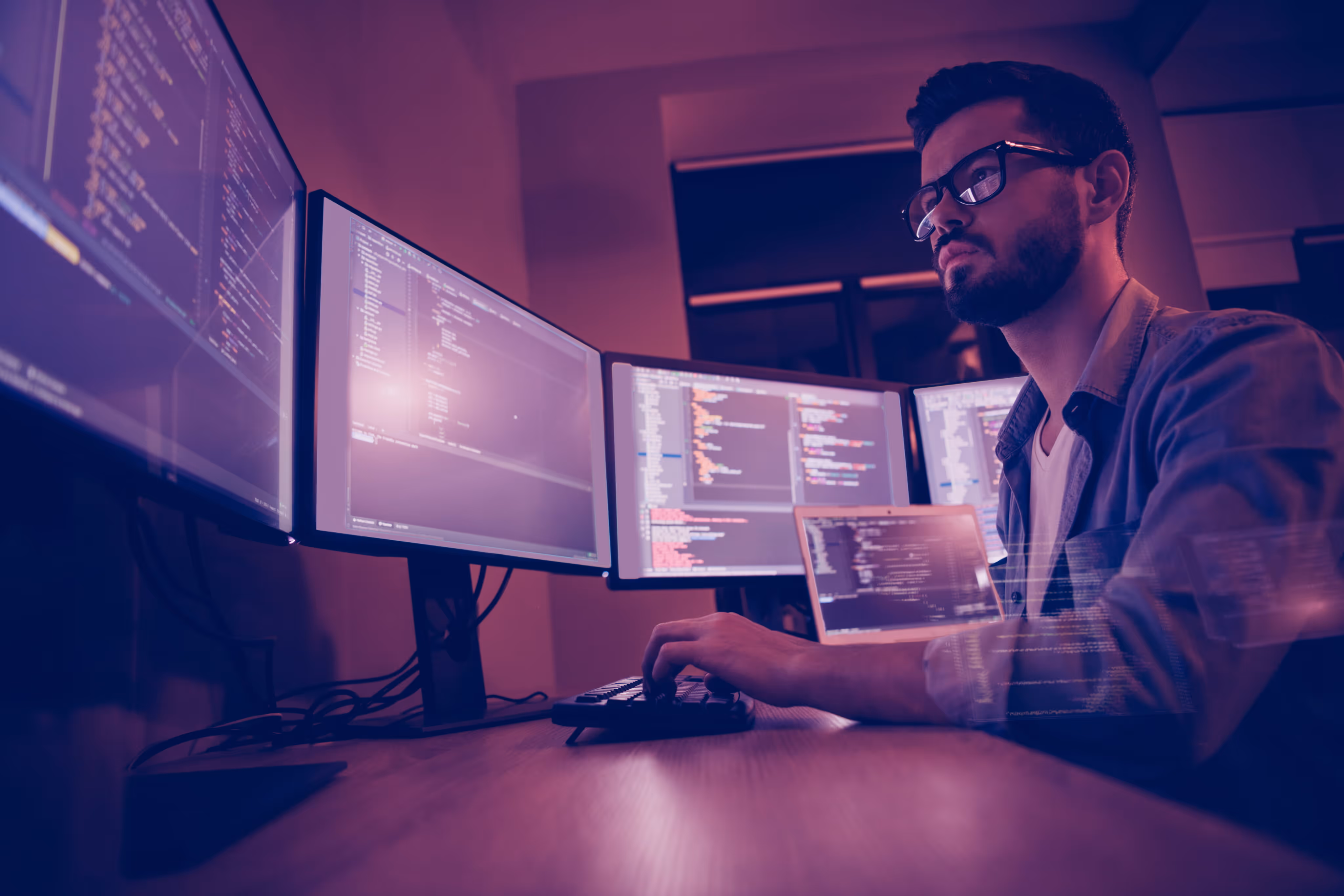Man with glasses programming on a keyboard with four monitors displaying code in a dimly lit room.