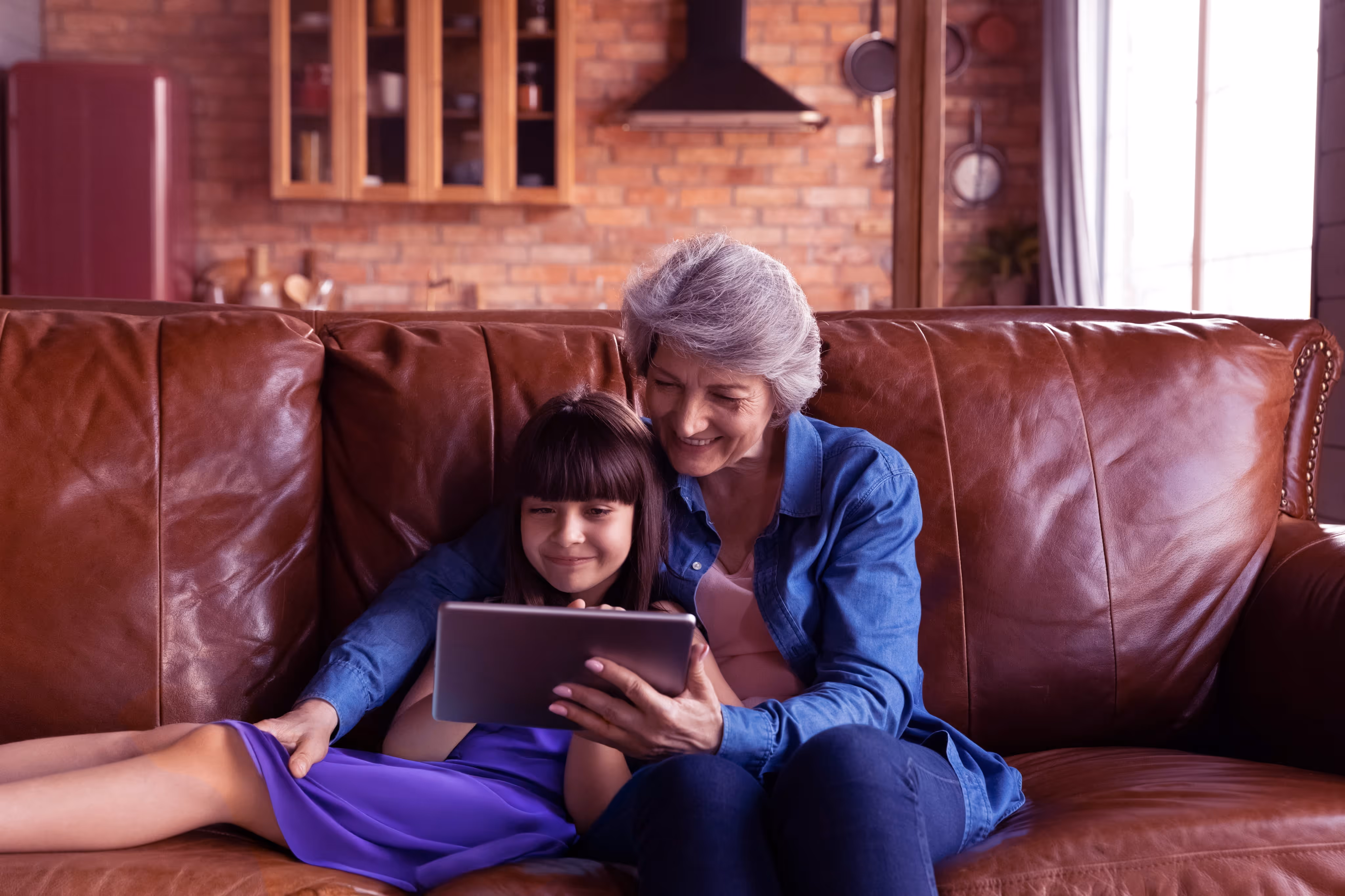 Grandmother and granddaughter sitting on leather couch watching a tablet together and smiling.