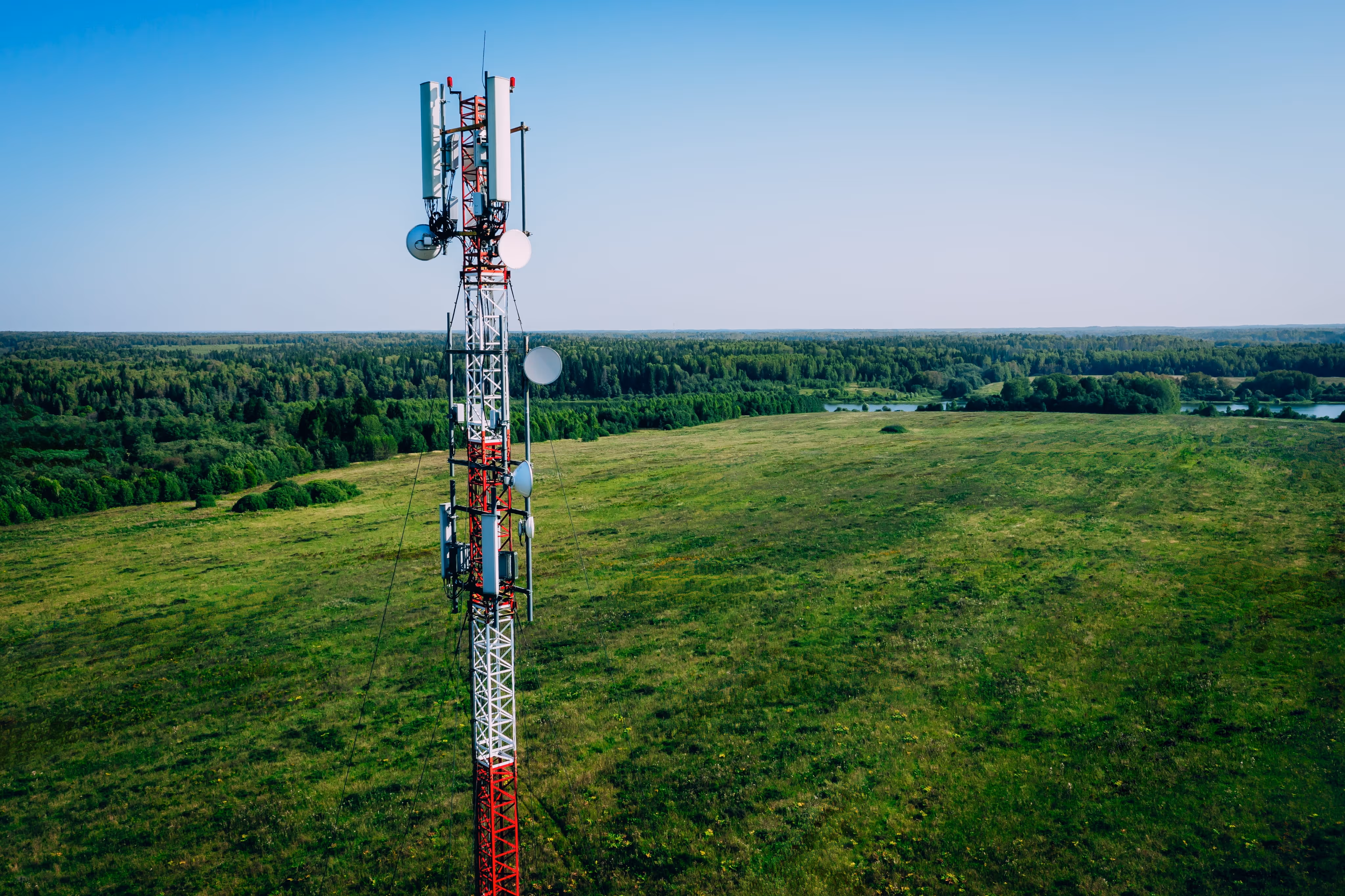 A red and white telecommunications tower stands in a green field with forest in the background.