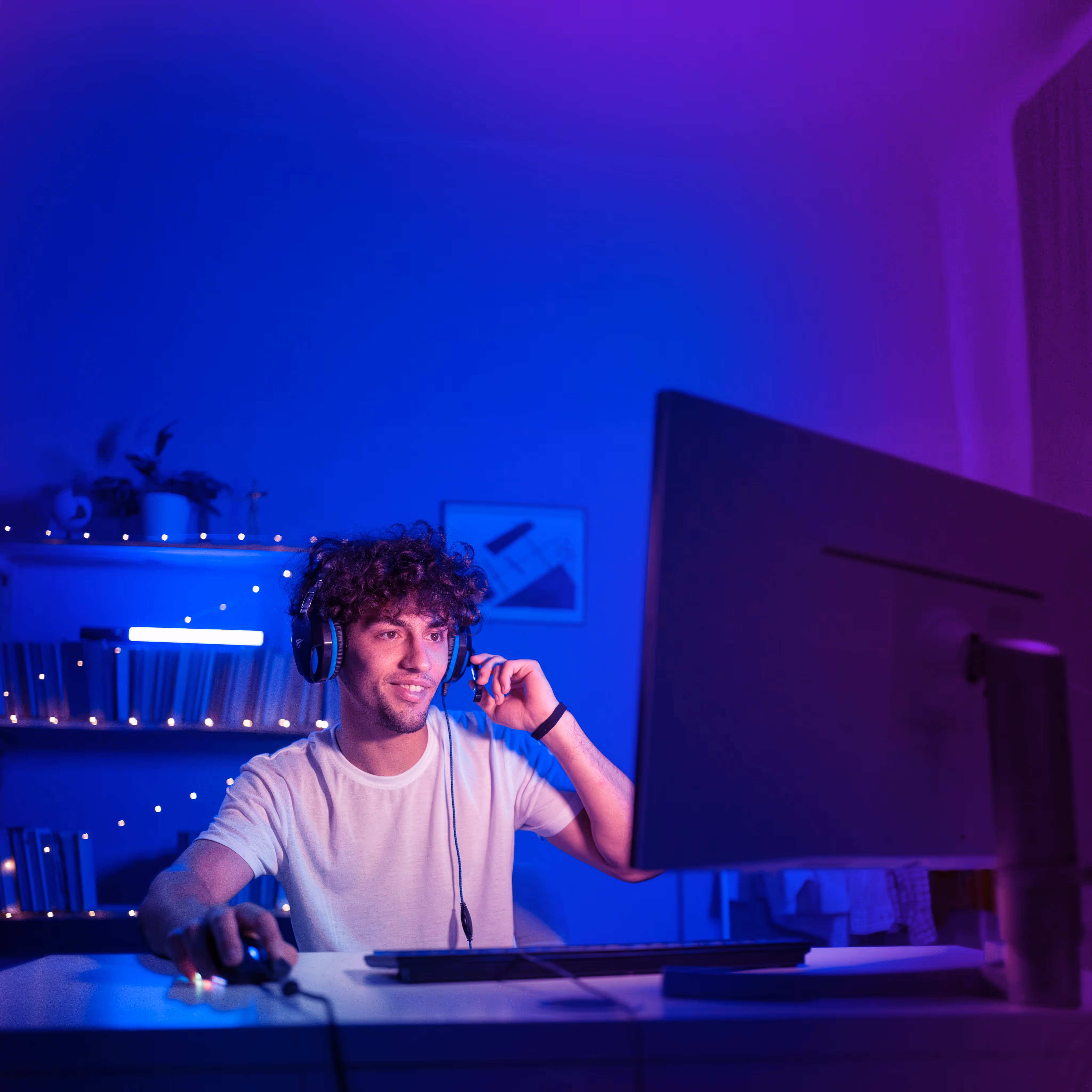 Young man with curly hair wearing headset and white shirt using computer mouse in blue-purple lit room.