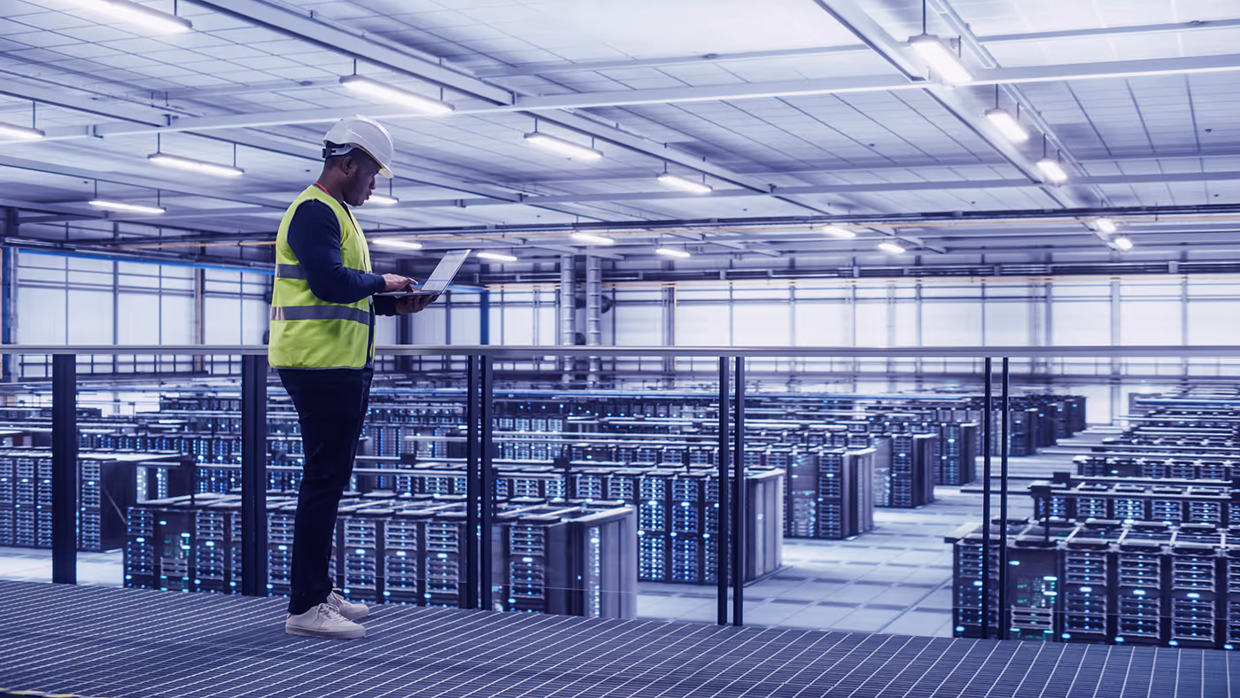 Technician in hard hat and safety vest using laptop in a data center with servers in the background.