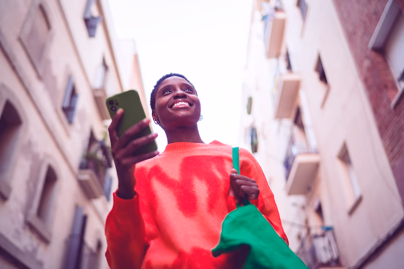 Smiling person in red sweater holding a green phone and tote bag walking outdoors in a city street.