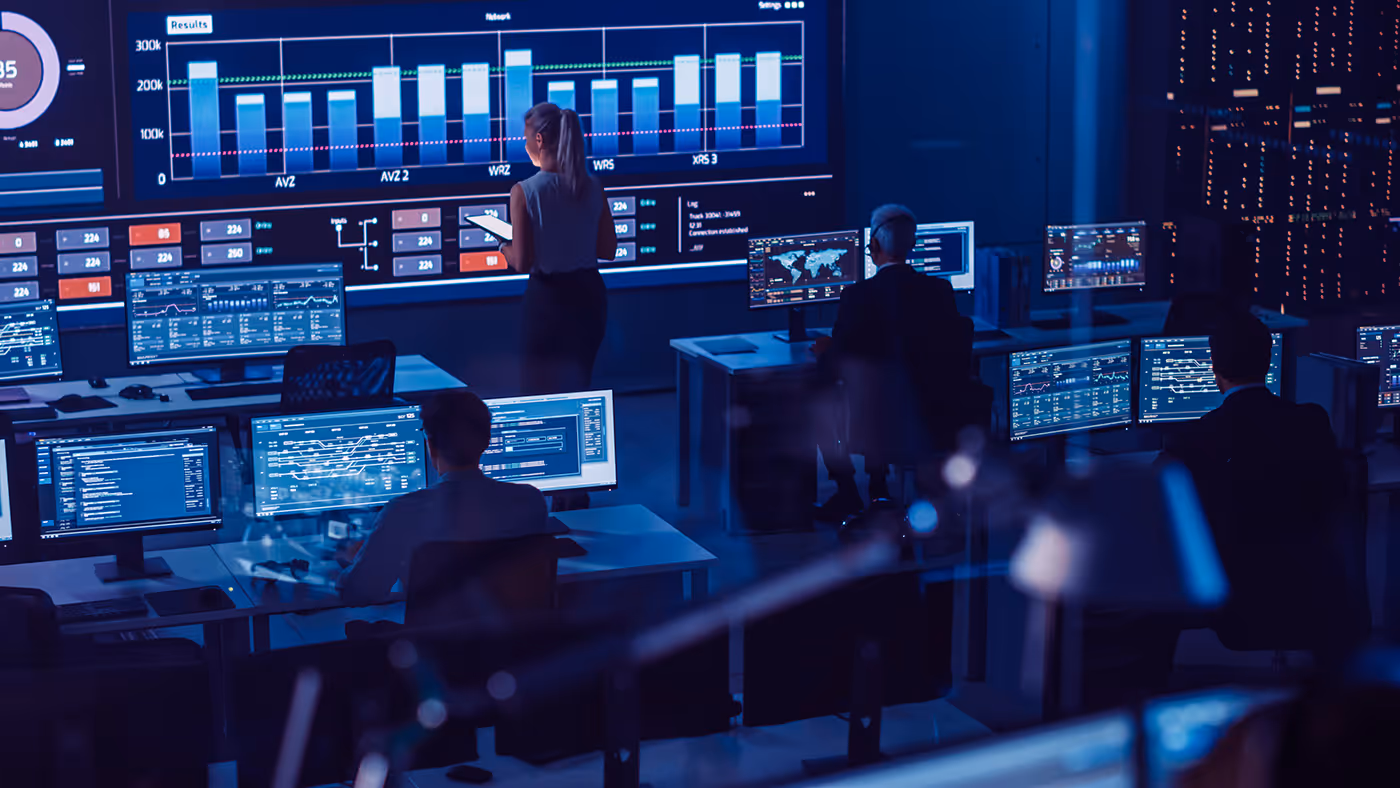 People working at computers in a dark control room with large data charts on the walls.