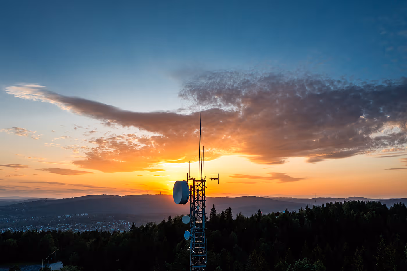 Communication tower with antennas against a sunset sky over forested hills and a distant town.