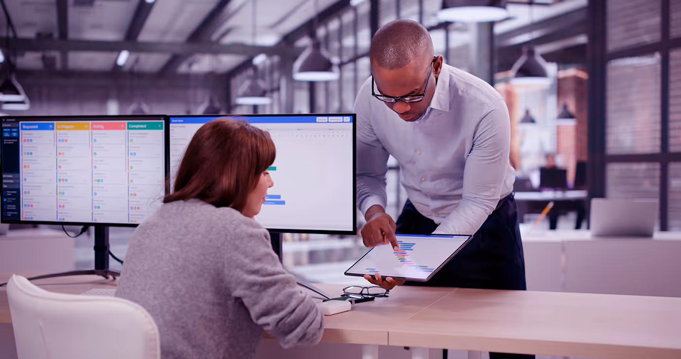 Man showing a woman a project chart on a tablet in an office with dual computer monitors.
