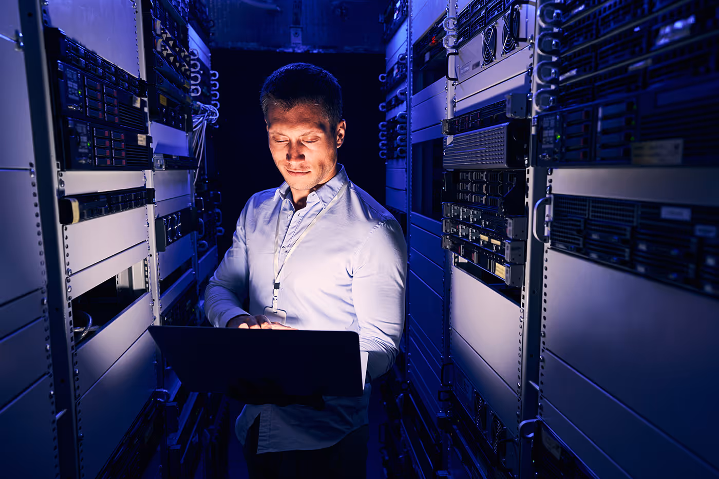 Man in white shirt working on laptop inside a dimly lit server room with racks of equipment.