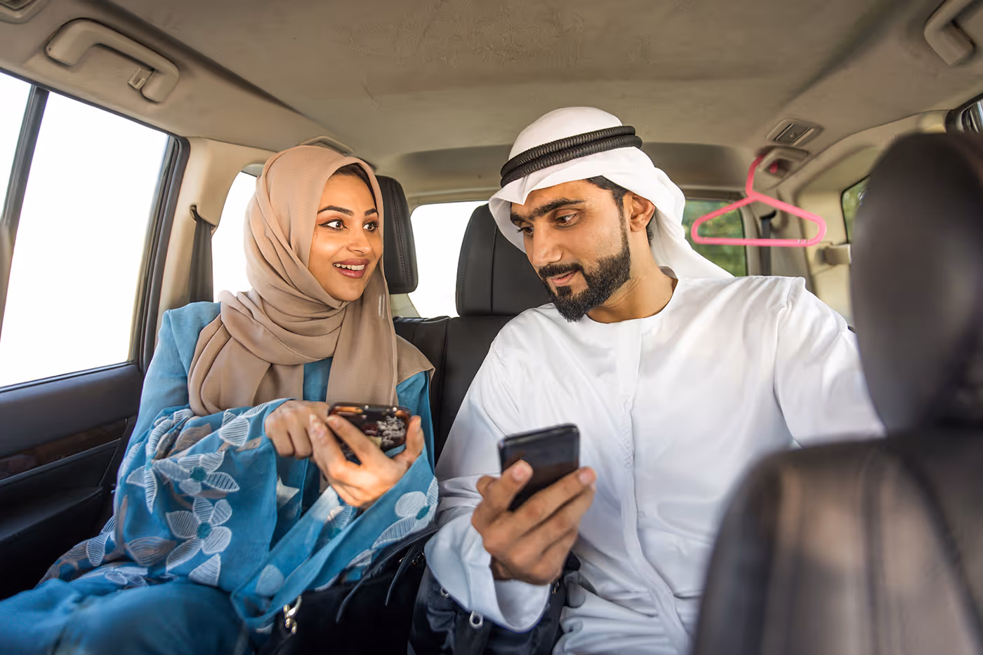 Arab man and woman in traditional dress sitting in a car, looking at phones and smiling.