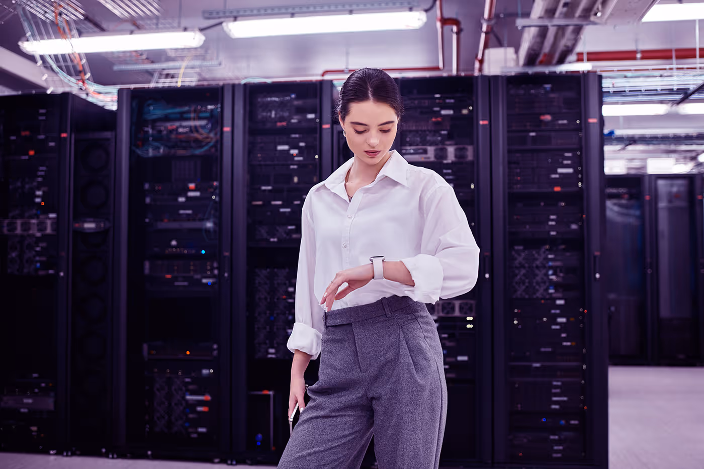 Woman in white shirt and gray pants checking smartwatch in a server room with racks.
