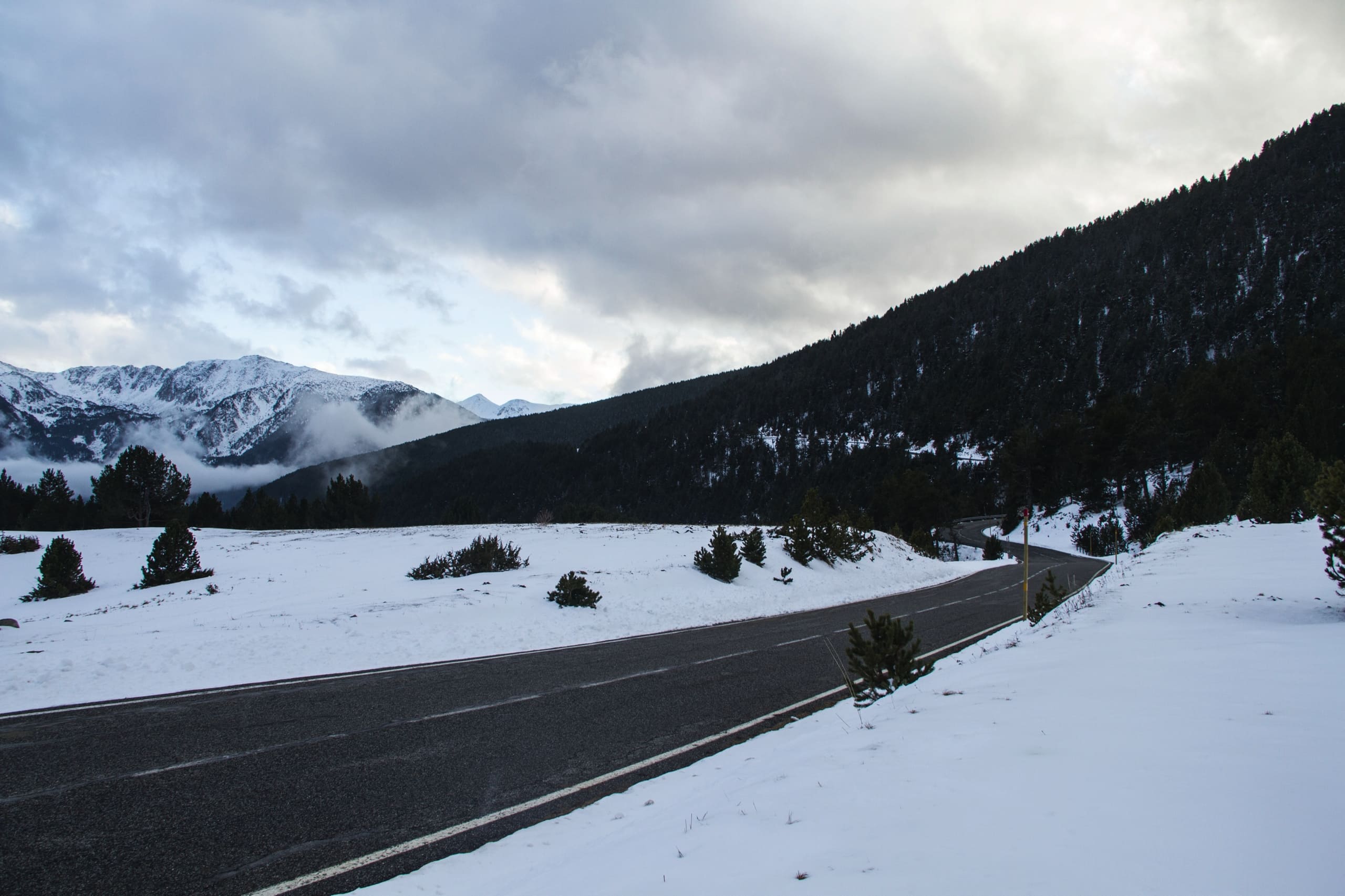 Snow-covered landscape with winding road, pine trees, and mountains under a cloudy sky.
