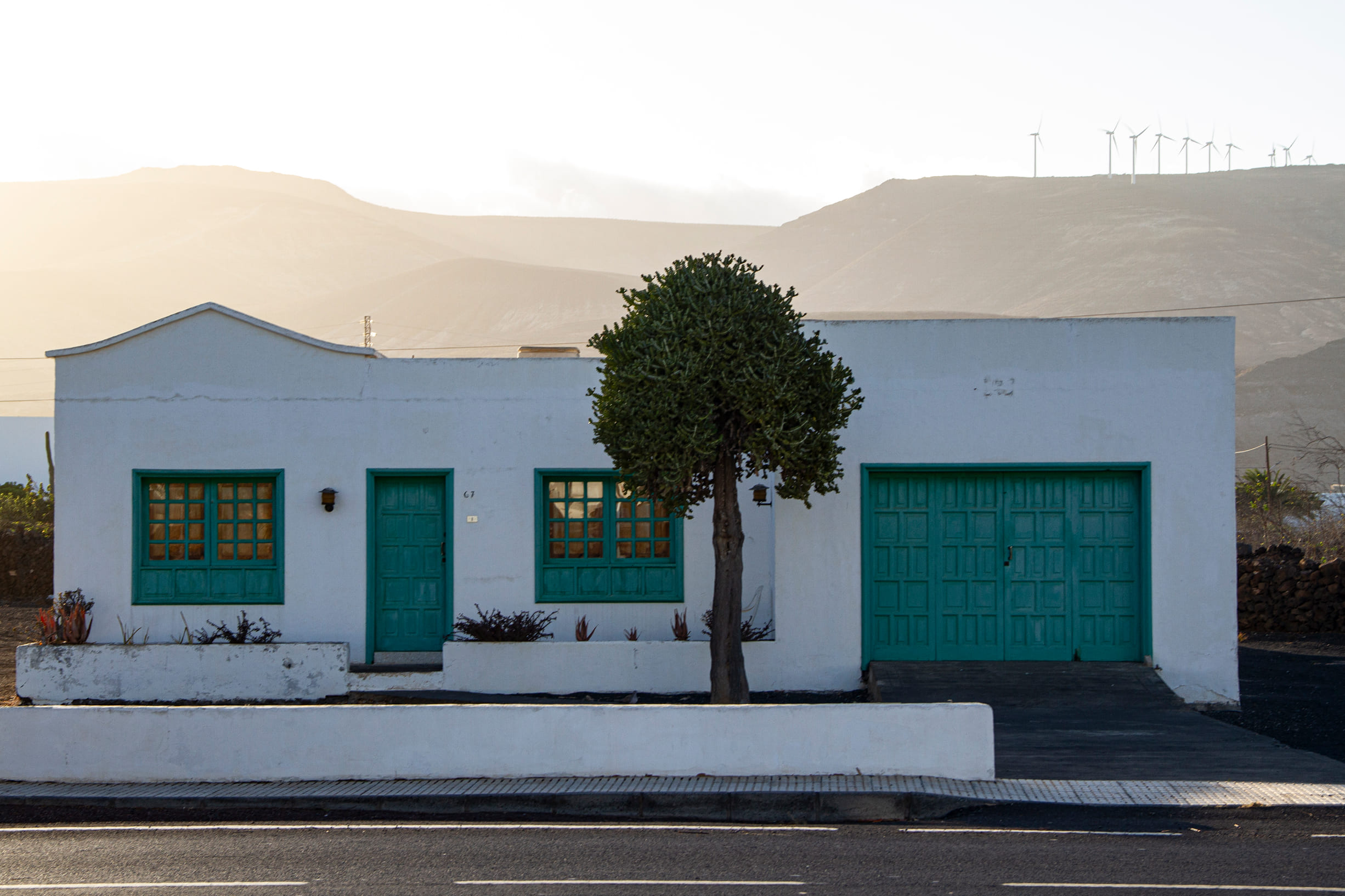 White house with turquoise doors and windows, a tree in front, and wind turbines on distant hills at sunset.