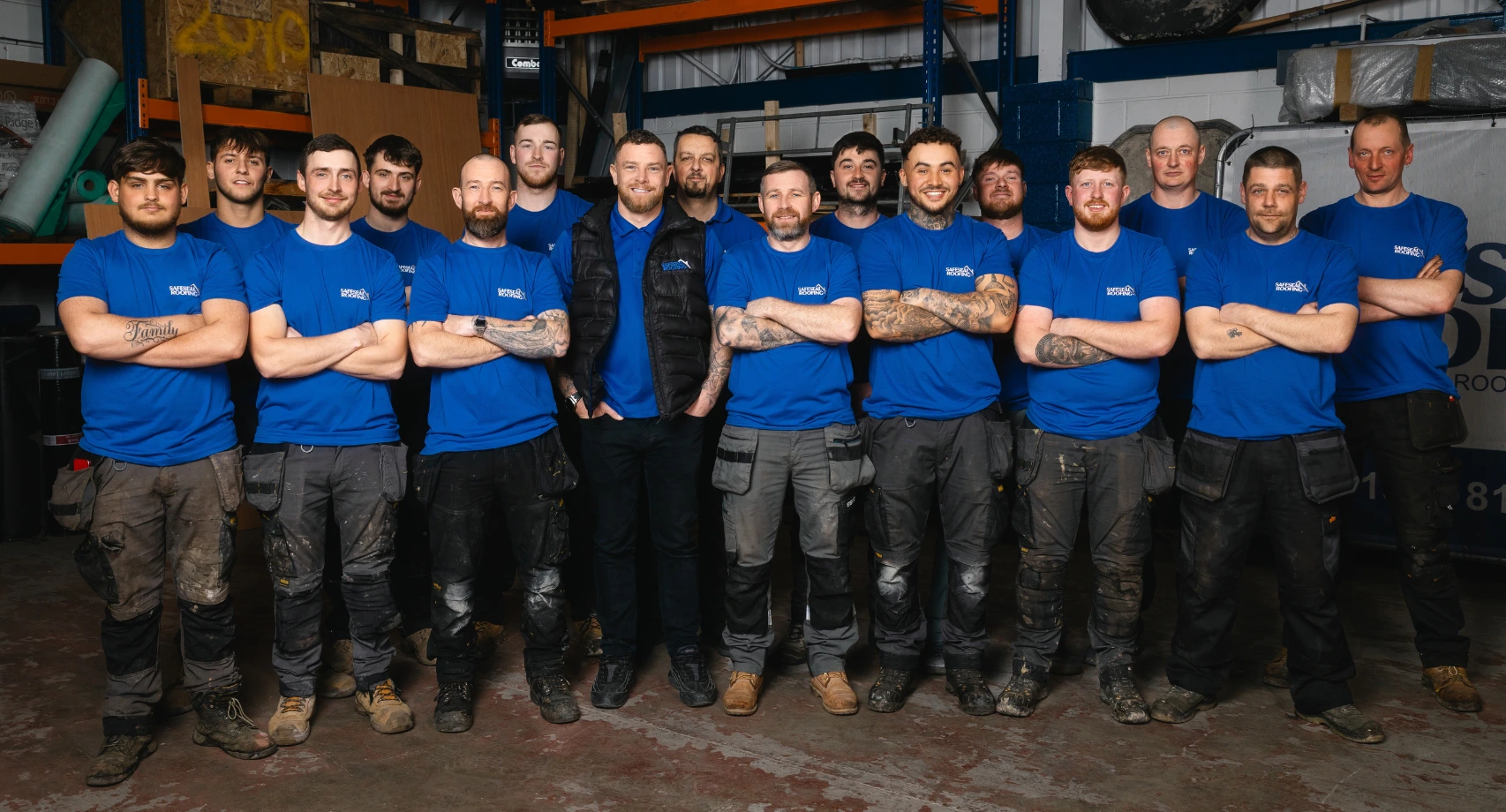 Group of 15 men in blue SafeSeal Roofing shirts standing with arms crossed in a warehouse.