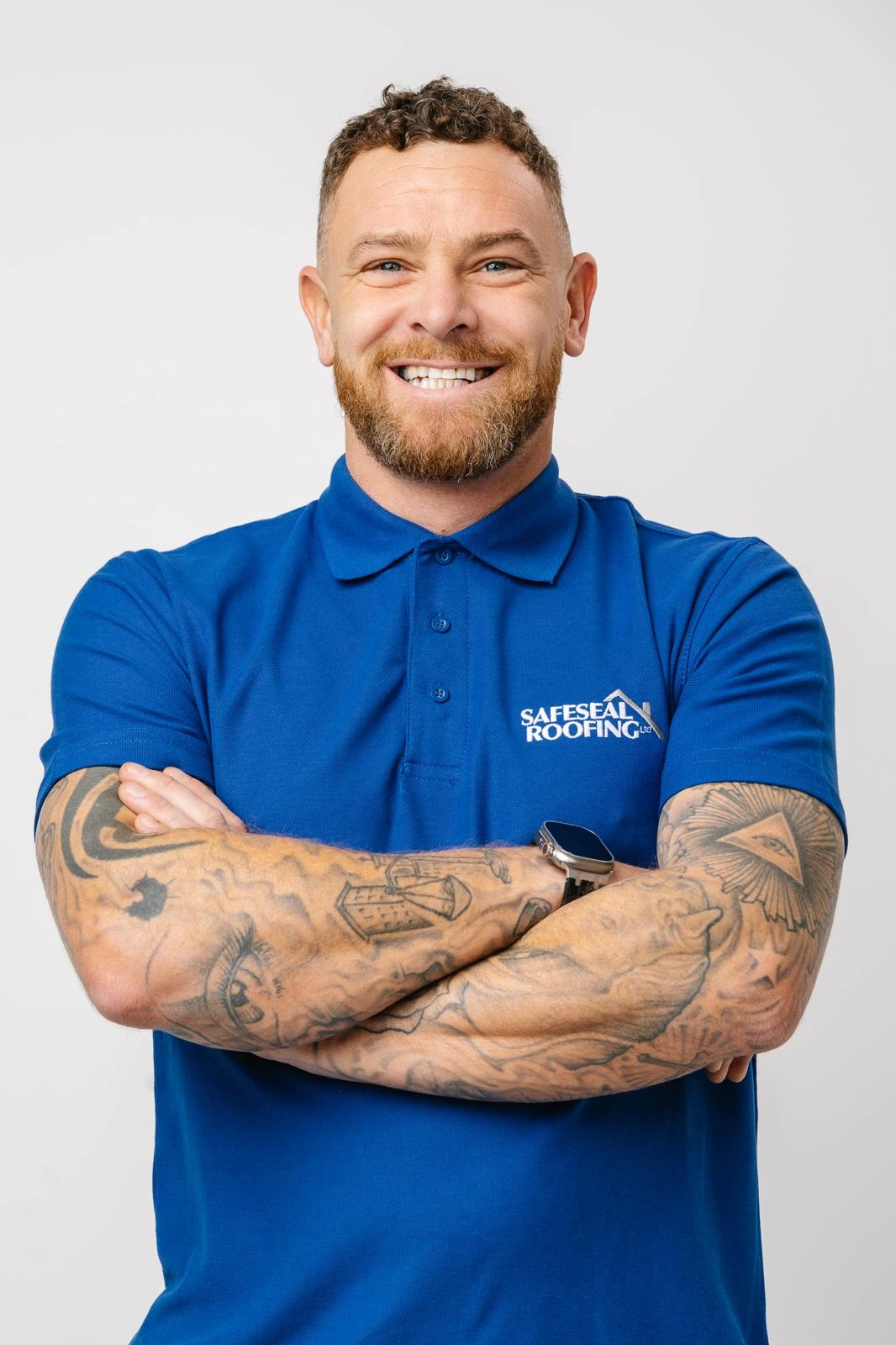 Smiling man with short curly hair, beard, and tattooed arms, wearing a blue Safeseal Roofing polo shirt.
