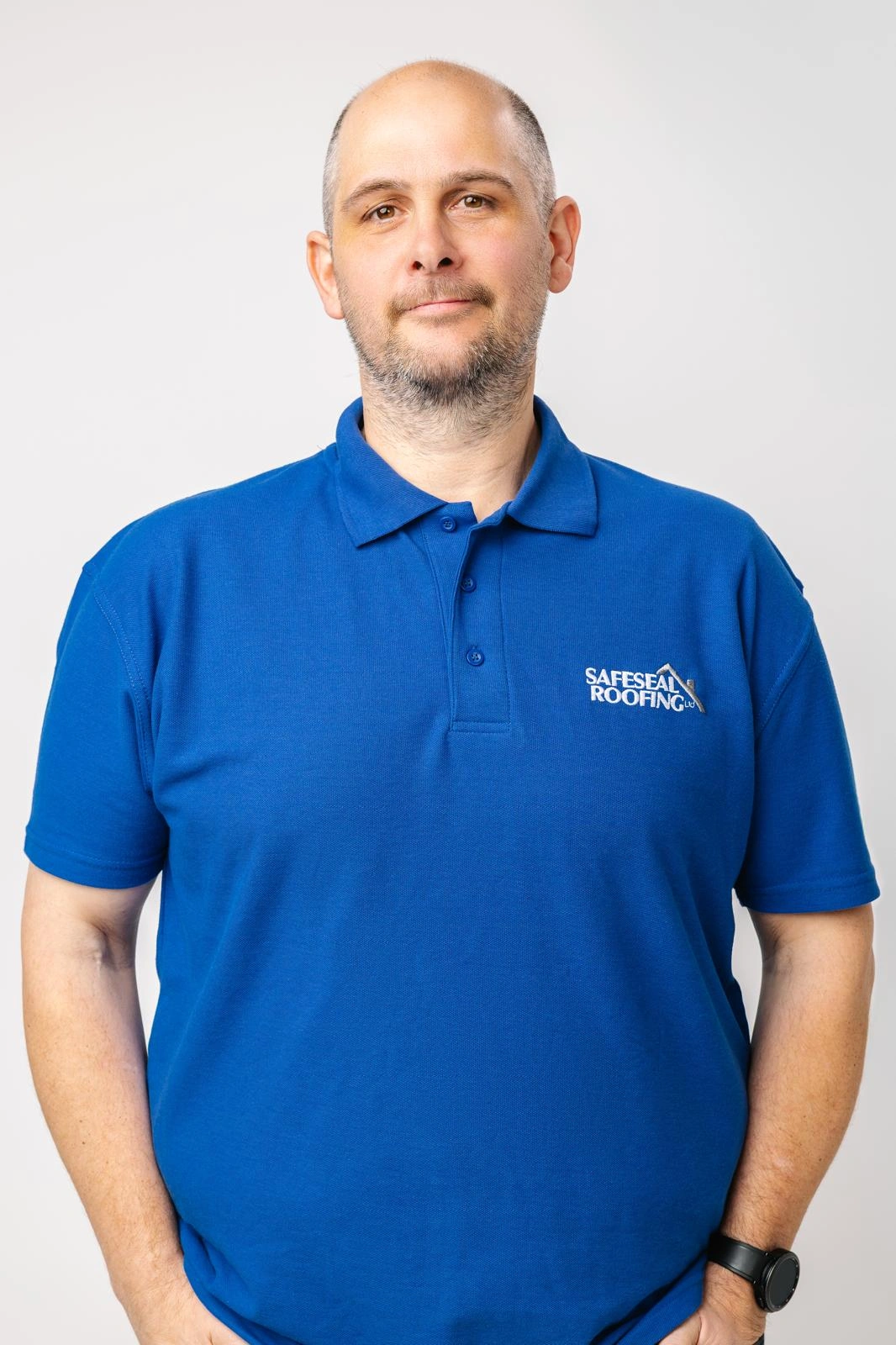 Man wearing a blue SafeSeal Roofing polo shirt standing against a light gray background.