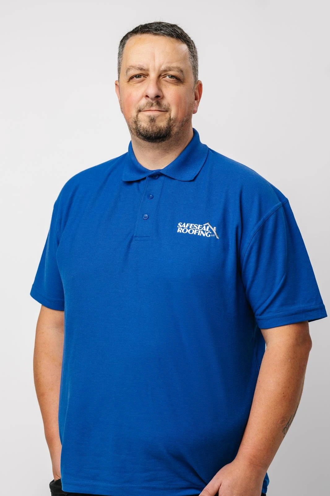 Man with short hair and goatee wearing a blue Safeseal Roofing polo shirt standing against a light gray background.