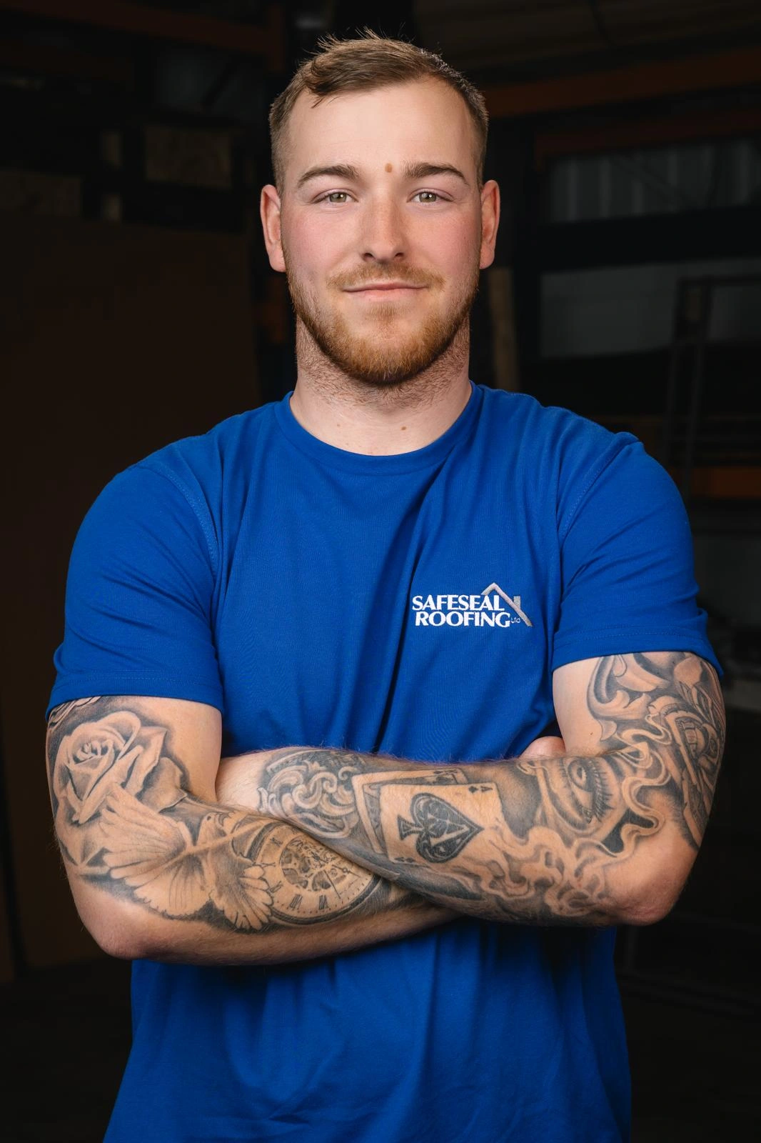 Man with short hair and beard wearing a blue Safeseal Roofing t-shirt, arms crossed showing detailed tattoos including a rose, dove, clock, and playing card.