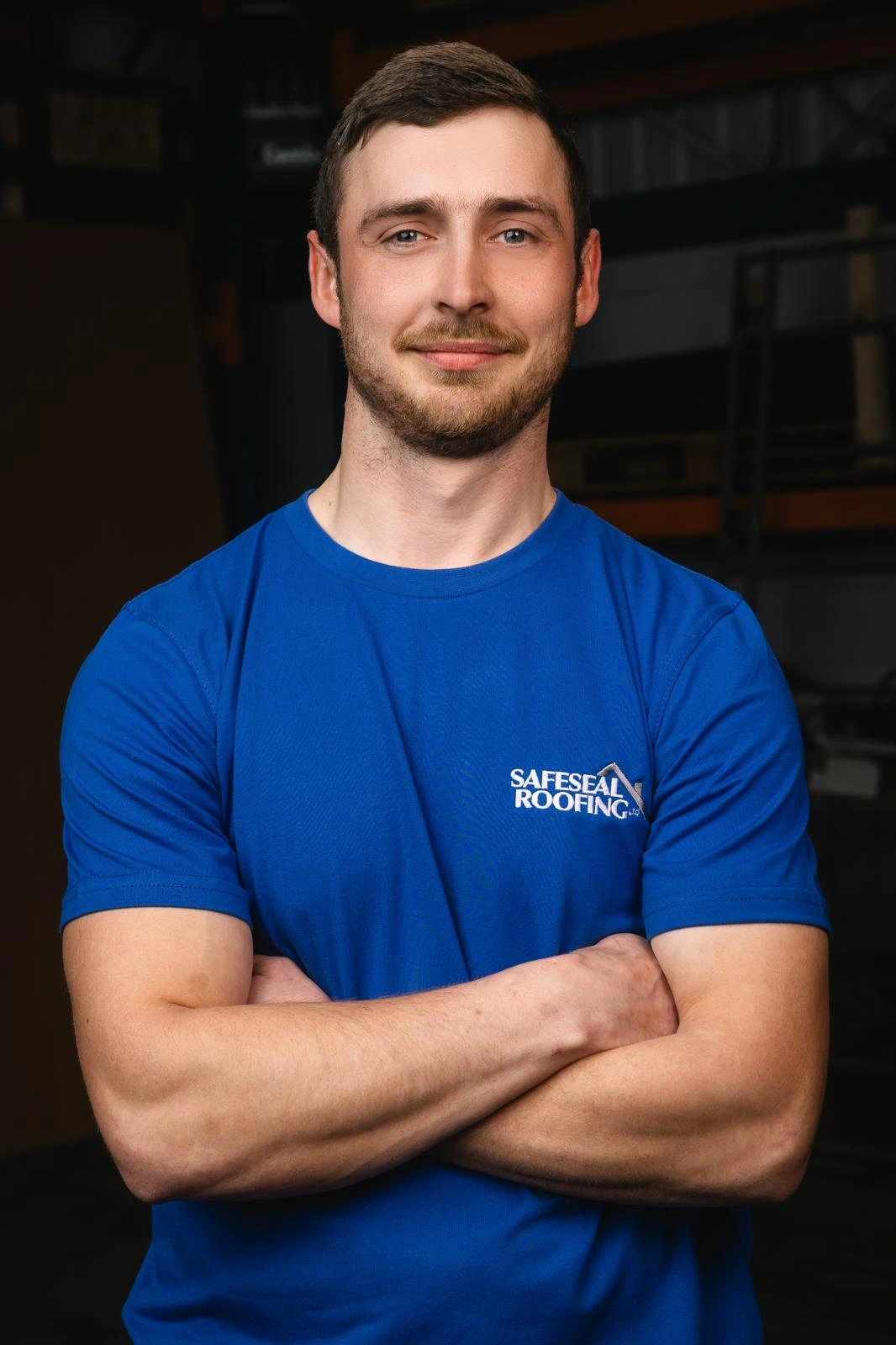 Man with short brown hair and beard wearing a blue Safeseal Roofing t-shirt with arms crossed, smiling slightly.