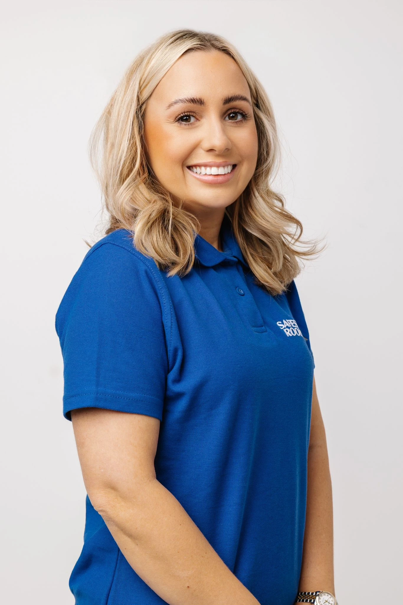 Smiling woman with blonde hair wearing a blue polo shirt with logo and a wristwatch.