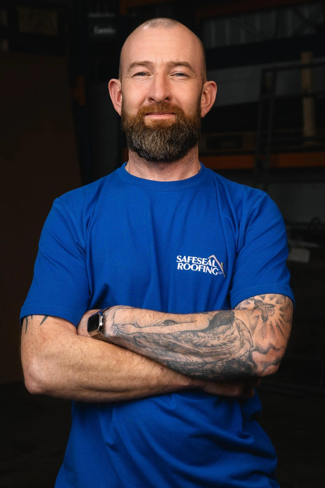 Bearded man with a tattooed arm wearing a blue Safeseal Roofing t-shirt with arms crossed.