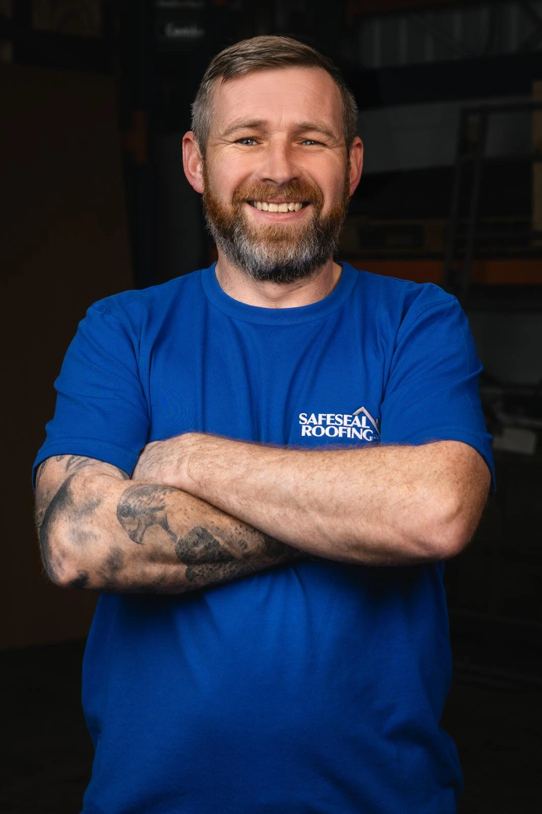 Smiling bearded man with crossed arms and tattoos wearing a blue Safeseal Roofing t-shirt.