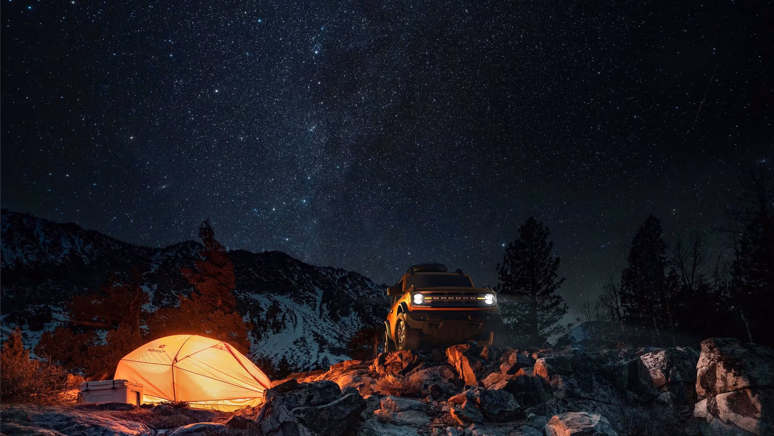 A yellow Ford Bronco driving on rocky terrain at night with a starry sky and a orange lit up tent.