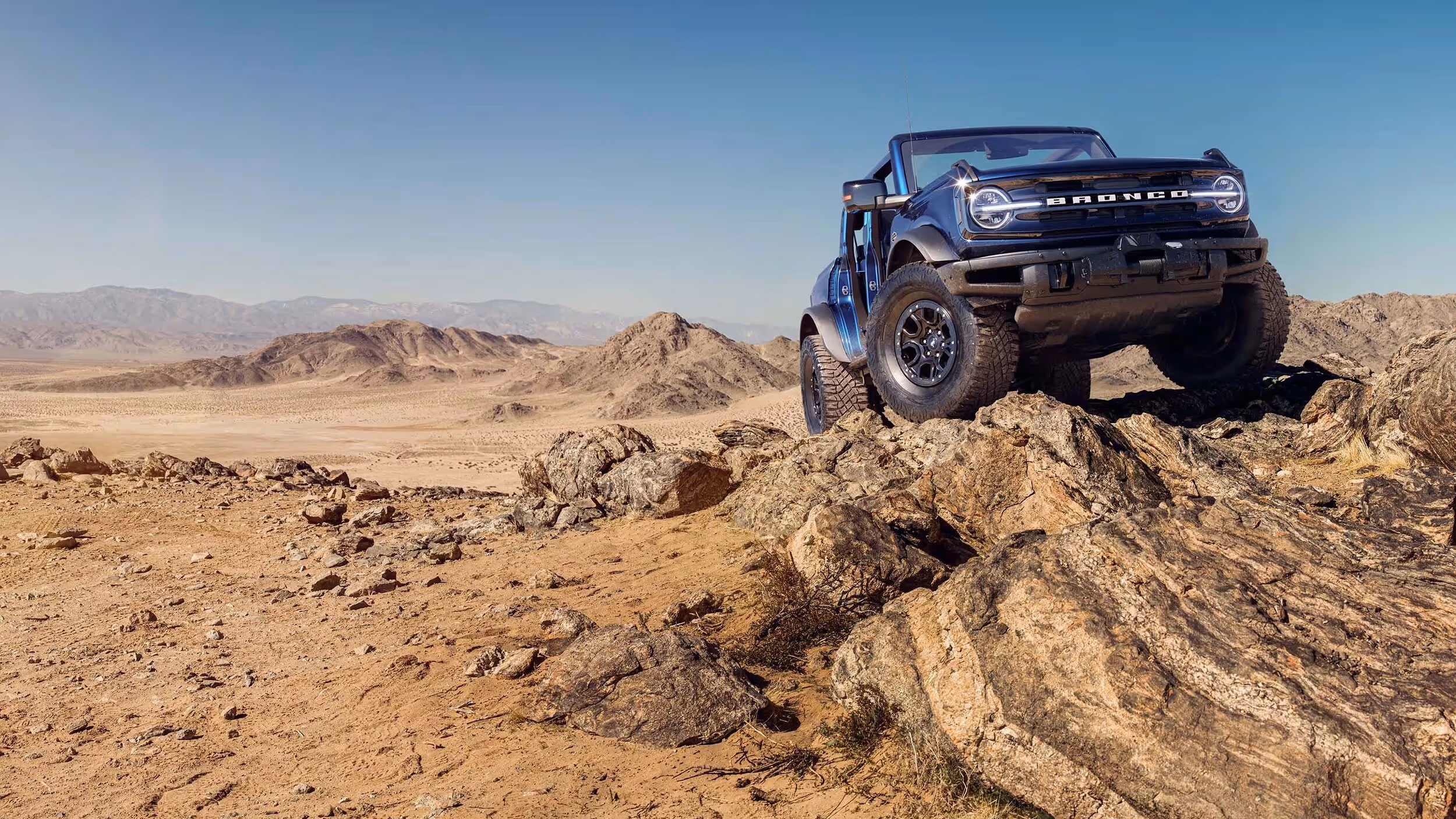Ford Bronco climbing over sand dunes under bright desert sun.