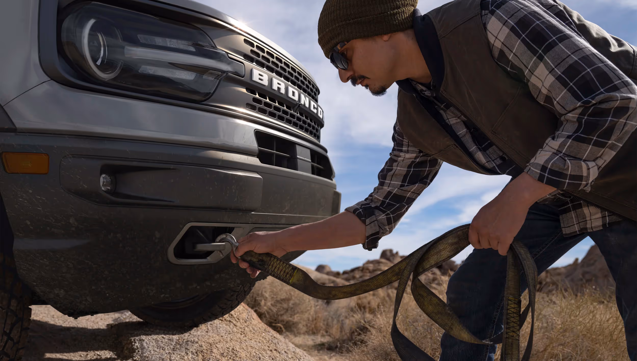 Person attaching tow strap to front of Ford Bronco Sport during off-road recovery.