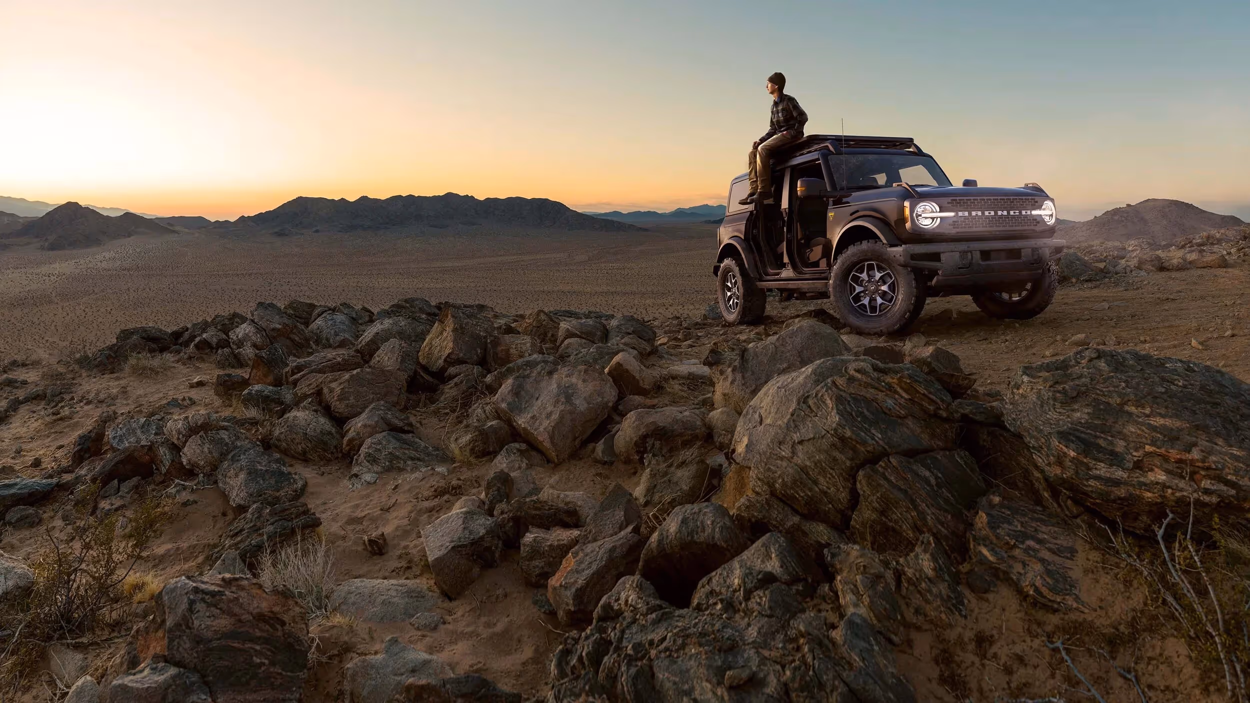 Lone adventurer standing beside Ford Bronco at golden hour.