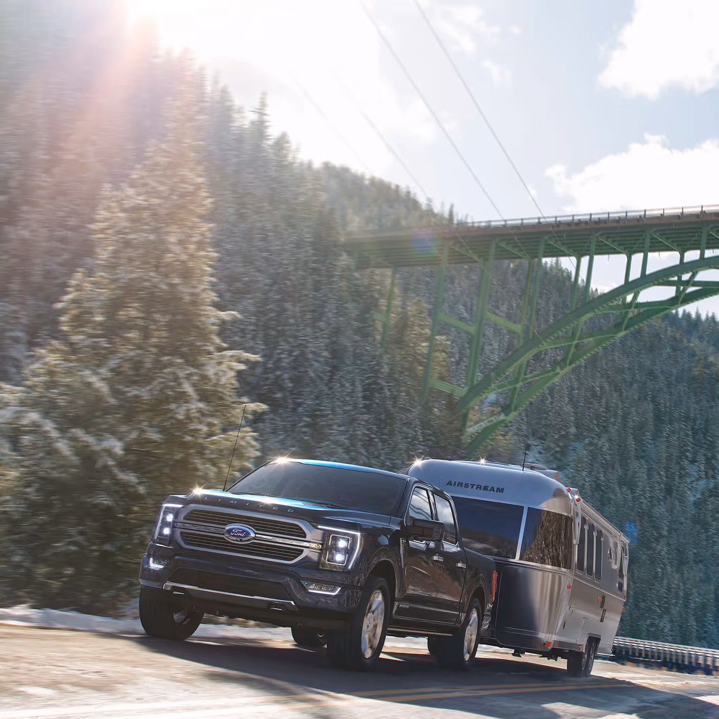 Ford F-150 towing an Airstream trailer across a mountain road beneath a steel bridge, sunlight breaking through clouds.