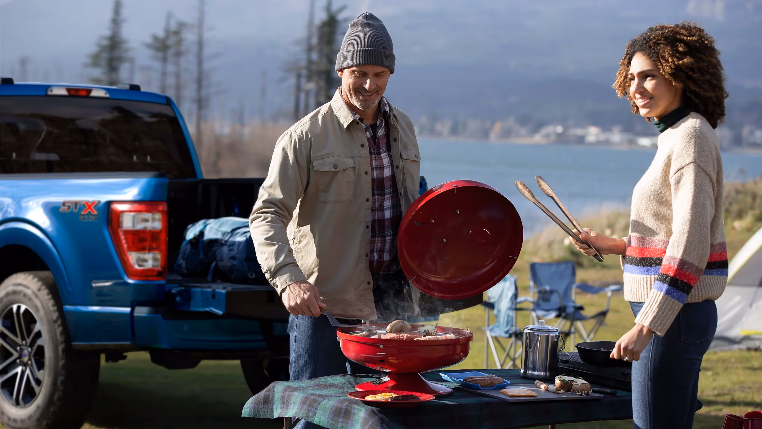 Couple cooking on a portable grill beside a blue Ford F-150 and Airstream trailer at a campsite.