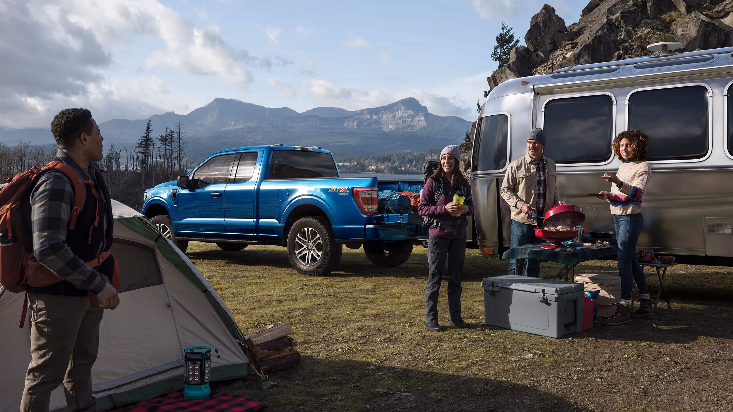 Blue Ford F-150 next to an Airstream trailer at a lakeside campsite with family gathered around gear.