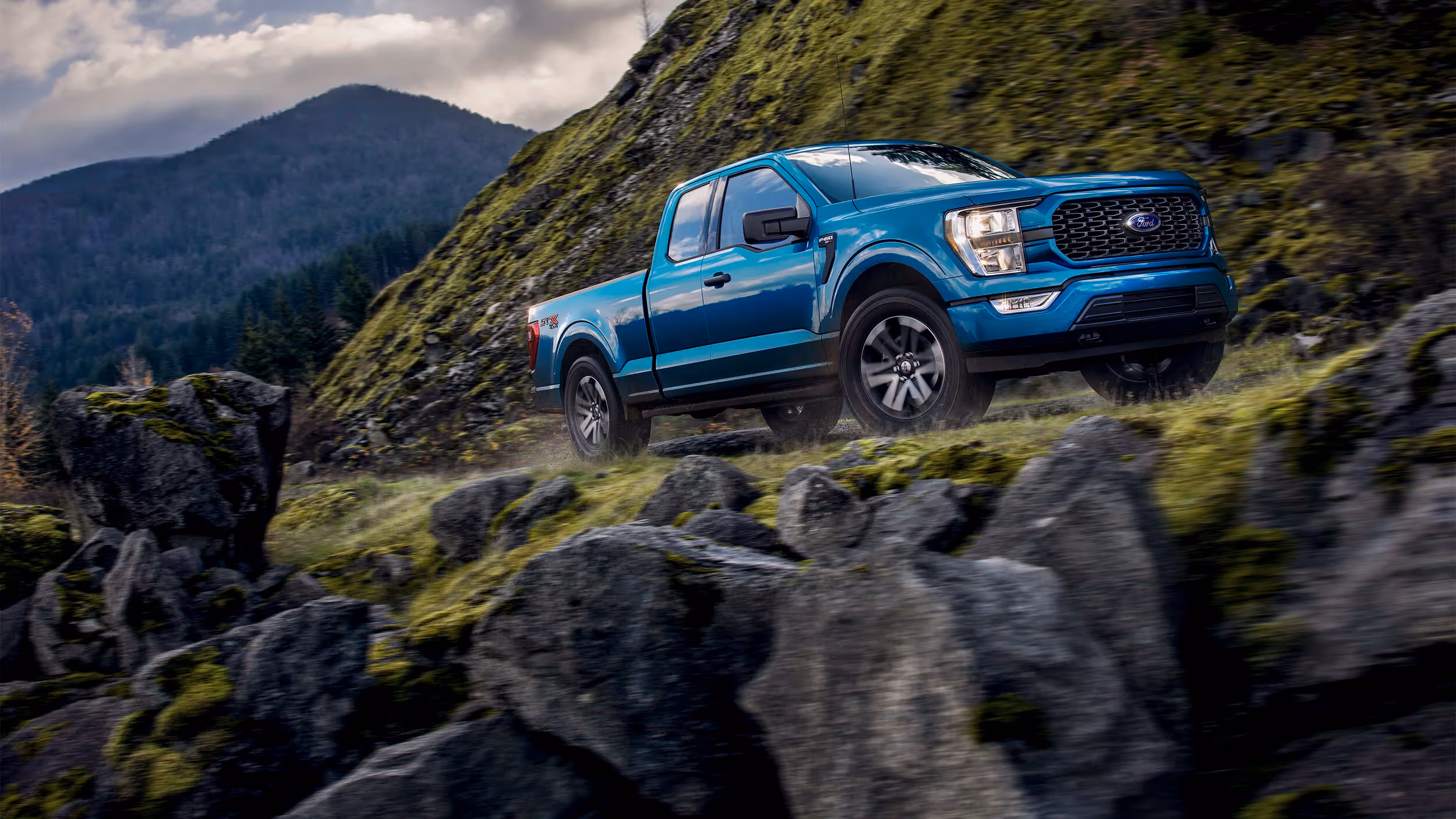 Blue Ford F-150 climbing rugged hillside terrain surrounded by pines and basalt boulders.
