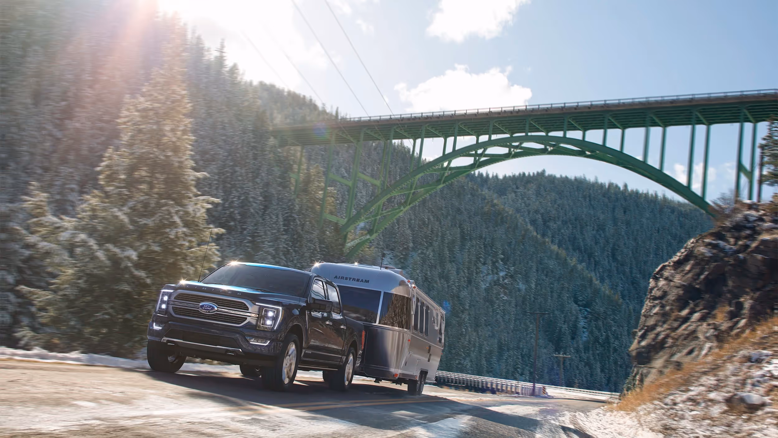 Ford F-150 towing an Airstream trailer across a mountain road beneath a steel bridge, sunlight breaking through clouds.