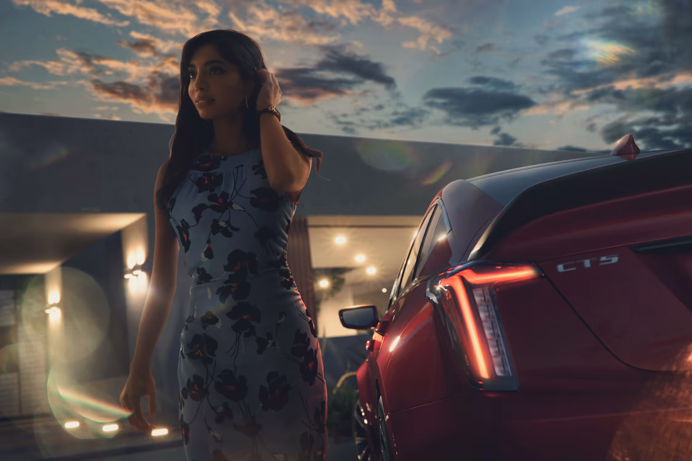 Woman standing beside Cadillac CT5 at dusk, warm sky and mountain backdrop behind her.