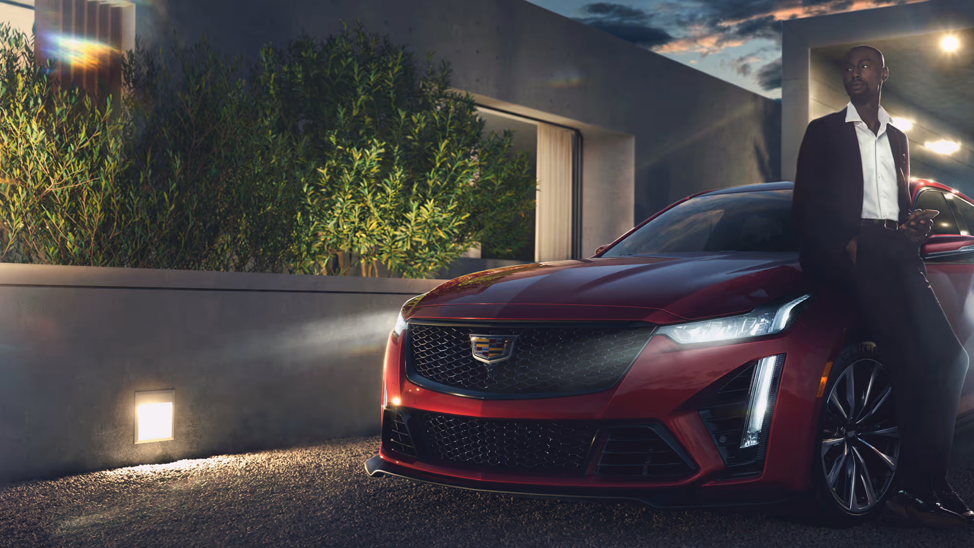 Man leaning on front of red Cadillac CT5 at sunset, house and greenery softly lit in background.