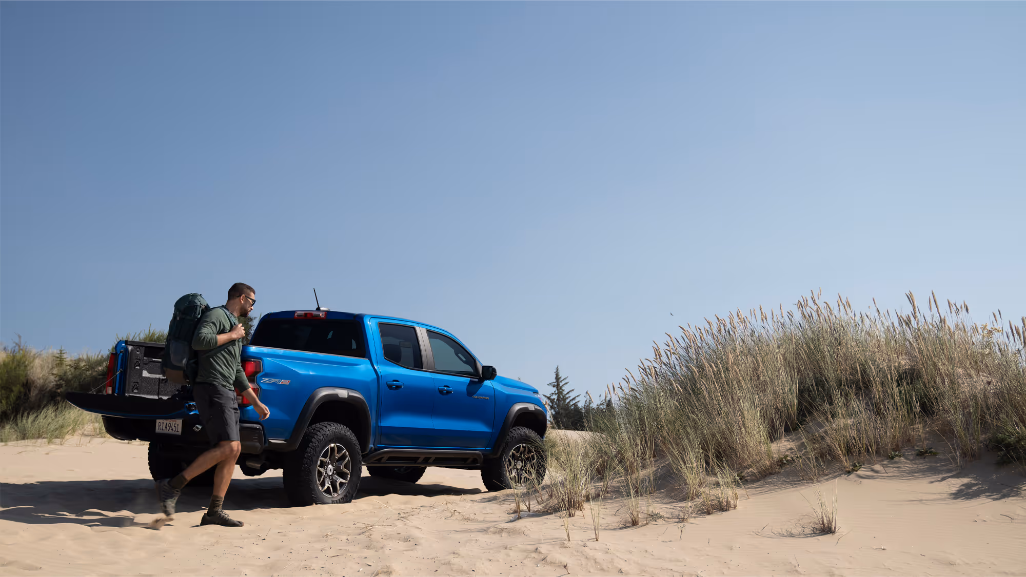 Man walking toward blue Chevy Colorado ZR2 with backpack, coastal grass in background.